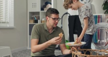 Bir adam ve oğlu çamaşır odasında oturmuş, çamaşırları sıraladıktan sonra dinleniyor, öğle yemeğinde peynirli tost yiyorlar..