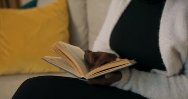 Close-up of girl of African appearance holding book in blue cover, reading and running finger along the lines. Girl is dressed in white cardigan, sitting on sofa on which lie yellow and blue pillows.