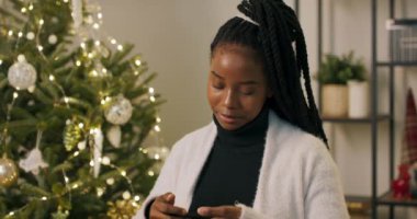 Siyah golf ve beyaz hırka giymiş Afrikalı görünümlü genç bir kız. Telefonu elinde tutuyor ve en iyi arkadaşını arıyor, iletişim kuruyor ve süslü Noel ağacını gösteriyor..