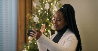 Afrika görünümlü güzel genç kız oturma odasında duruyor ve elinde telefonla kız kardeşini arıyor. Kız görüntülü konuşma yapıyor, gülümsüyor ve Noel ağacını gösteriyor..
