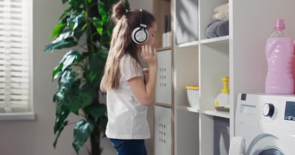 Smiling Little Girl Wearing Headphones Listening Music Enters Laundry Room — Wideo stockowe