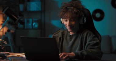 Kıvırcık saçlı genç bir çocuk uzaktan öğrenmeyle mutludur. Bir öğrenci kablosuz kulaklıklı bir bilgisayarın önünde oturuyor ve bir öğretmenin dersini dinliyor. Dersler internet üzerinden yapılıyor..