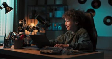 Young talented gifted boy sits in room in the dark and programs a robot on laptop, adds new features, high-fives it, interest in robotics technology.