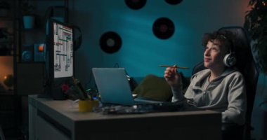 Bir çocuk akşam odasında bilgisayar ekranı ve dizüstü bilgisayarın önünde oturur, bir okul projesi üzerinde çalışır, kalemle bir defter çizer, çalışmalarını planlar, düşünür..