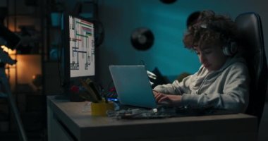 Bir çocuk teknoloji sınıfı için bir bina tasarlıyor, ödev yapıyor, defterine kurşun kalemle çiziyor, grafiklerle dolu bir bilgisayar ekranına bakıyor. Masası olan bir erkek odası, duvarında plaklar var..