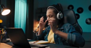 Teenager studies diligently in front of laptop, takes notes with pencil in notebook, rewrites, listens to music on wireless headphones sings smiles.