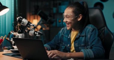 Young talented gifted girl sits in her room in the dark and programs a robot on her laptop, adds new features, high-fives it, interest in robotics technology.