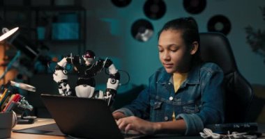 Cheerful, intelligent student sitting at desk at home constructing robotic device in spare time, child smiles develops hobby.
