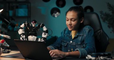 Cheerful, intelligent student sitting at desk at home constructing robotic device in spare time, child smiles develops hobby.