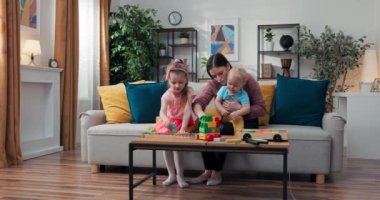 A woman sits watching two children at home. Caregiver entertains toddlers with toys, shows blocks to youngest boy, girl in pink dress plays dulcimer, babysitting.