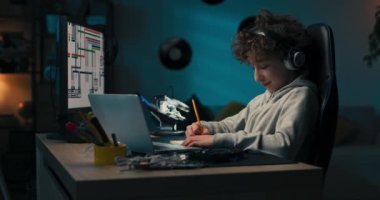 A boy sits in room in the evening in front of computer screen and laptop, working on a school project, drawing in a notebook with a pencil, planning the look of his work, thinking.