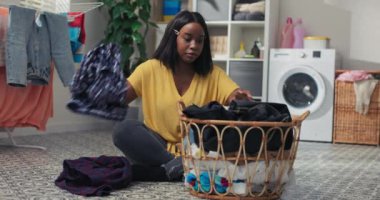 Genç bir anne çamaşır odasında yerde otururken ev işi yapar. Kadın çamaşırları katlar, giysileri ayırır, banyoyu temizler..