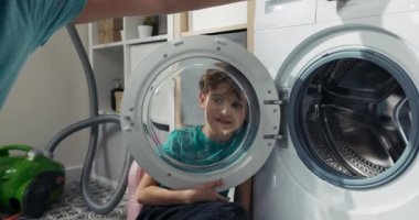 Siyah gözlü gülümseyen çocuk çamaşır makinesinin yuvarlak cam kapısından bakar, ev işlerini yaparken aylak aylak dolaşır..