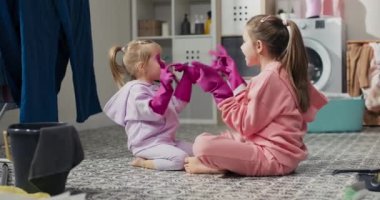 Two sisters play together while cleaning the apartment sit on the bathroom floor, fool around with a battle of pink rubber protective gloves.