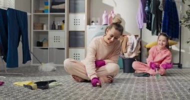 Concept of cleaning at home with child. Mom sits on bathroom floor in laundry room and she and daughters scrub floor with a brush and liquid detergent. Teaching children to clean.
