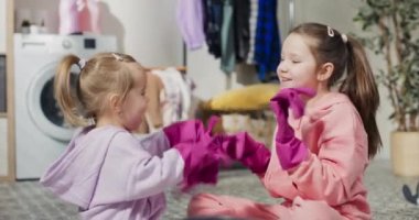 Two sisters play together while cleaning the apartment sit on the bathroom floor, fool around with a battle of pink rubber protective gloves.