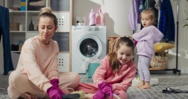Happy family cleaning house together. Twin sisters helping mommy to tidy up apartment. Portrait of young mother and cute little daughters smiling while washing the floor at home.