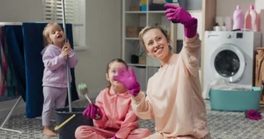 Gülümseyen bir kadın telefonda bir videonun önünde konuşuyor. Kızların evi temizlerken kıza yardım ettiğini gösteriyor. Yerleri paspaslarken çocuklarla sosyal medyada selfie.