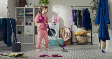 Gülümseyen mutlu bir çocuk çamaşırhanede ev işleri yaparak vakit geçirir. Pembe eşofmanlı bir kız paspasla yerleri yıkıyor..