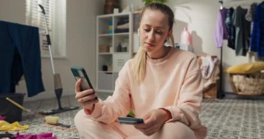 A young woman noticed a lack of clean supplies while cleaning. She orders groceries via an app on phone, pays with a card and waits happily for delivery.