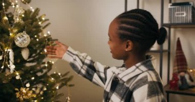 Afrikalı görünümlü genç bir kız Noel ağacı süslemiş. O, çelenkler ve süslemeler astı. Kız Noel ağacına hayran ve çelengi tamir ediyor. Tatmin olmuş durumda..