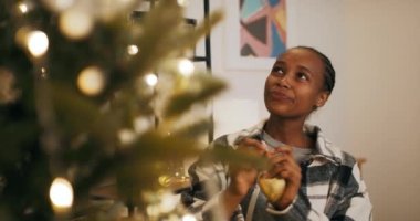 Noel arifesinin büyüsü. Afrika görünümlü çekici genç kız Noel ağacını süslüyor. Kız kalp şeklindeki süsü tutuyor ve asıyor. Işıklar yanıp sönüyor..