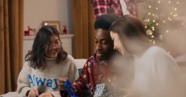 Oturma odasındaki kanepede oturan gülümseyen arkadaşlar. Asyalı kız, Afrikalı erkek ve elinde hediyeler olan esmer kız. Adam kutuyu salladı ve ortadakini yakalamaya çalıştı. O, hediyeyi açtı ve sevindi..