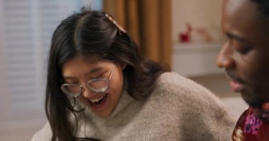 Christmas party. An Asian-looking girl with glasses received gift and unpacked it. The brunette is sincerely smiling and happy to receive gift. Friends sitting next to her are happy for friend.