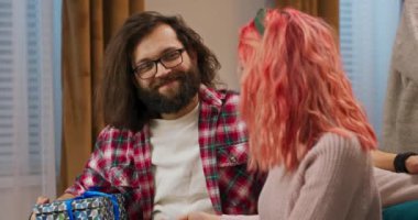 Christmas Eve. Friends sitting on the couch. A guy with beard with glasses holds a gift behind his back and gives it to a girl. The beauty with red curly hair is pleasantly surprised and hugs the guy.