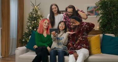 During the Christmas party, five friends gathered in the living room by the sofa and have video chat. Asian girl in glasses holds phone in hands. Friends communicate and laugh.