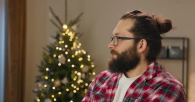 A young man with beard and glasses stands in the living room. The guy is looking for friends. When he found them, smile appeared on his face. The lights on the Christmas tree twinkle in the room.