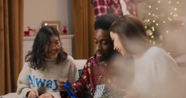 Oturma odasındaki kanepede oturan gülümseyen arkadaşlar. Asyalı kız, Afrikalı erkek ve elinde hediyeler olan esmer kız. Adam kutuyu salladı ve ortadakini yakalamaya çalıştı. O, hediyeyi açtı ve sevindi..