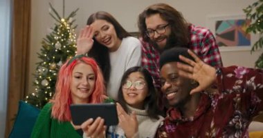 En iyi arkadaşlar aileleriyle görüntülü sohbet ederler. Gözlüklü Asyalı kız telefonu elinde tutuyor ve gülümsüyor. Erkekler ve kızlar mutlu bir şekilde iletişim kurar ve herkese mutlu Noeller diler..