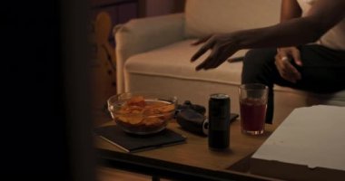 Gülümseyen adam oturma odasındaki kanepede dinleniyor, beyaz askılı afro gömlekli adama uzanıyor, uzaktan kumandayı tutuyor, müziği açıyor, kanalları açıyor, en sevdiği programı açıyor,.