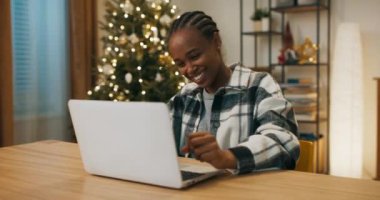 African-looking girl playing online game with friend on laptop. Ahead is the decisive round and the girl wins. She is infinitely happy and smiling. Behind shimmers Christmas tree.