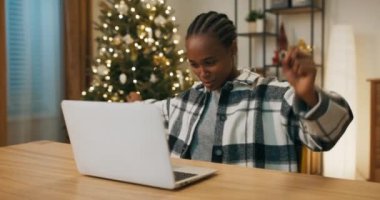 African-looking girl playing online game with friend on laptop. Ahead is the decisive round and the girl wins. She is infinitely happy and smiling. Behind shimmers Christmas tree.