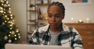 An African-looking girl is sitting at table in living room. She looks intently at the laptop screen and writes to co-workers. Together they are preparing project. A Christmas tree flickers in the room