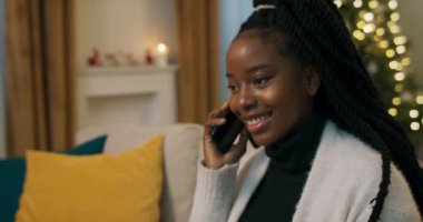 Cheerful girl of African appearance sits on the couch and talks on the phone with friends. They are planning trip for the Christmas holidays. The girl smiles and gestures with hands.