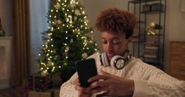 Kış sıcağı giymiş kıvırcık saçlı çocuk kanepede dinleniyor kablosuz kulaklıklarla podcast dinliyor, telefonda şarkılarını değiştiriyor, kahkahalar atıyor, arka planda Noel ağacı var..