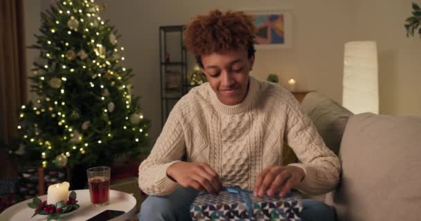 好奇心旺盛な男がクリスマスツリーの近くのソファに座っている 白いセーターを着た笑顔のハンサムな男がギフトボックスを開きます 彼は失望している 悲しい 彼は驚きが好きではない — ストック動画