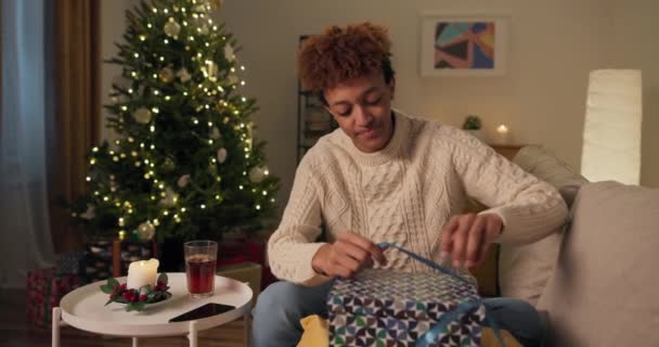 一个好奇的男人坐在圣诞树旁的沙发上 一个穿着白色毛衣 面带微笑的英俊男子打开礼品盒 他很失望 很伤心 不喜欢这个惊喜 — 图库视频影像
