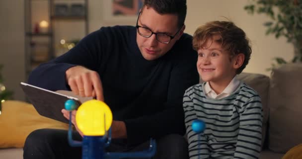 息子は父親と一緒にソファに座って惑星系男のモデルと遊んでいる彼は惑星を命名している例としておもちゃを使って宇宙の建設について少年に説明している — ストック動画