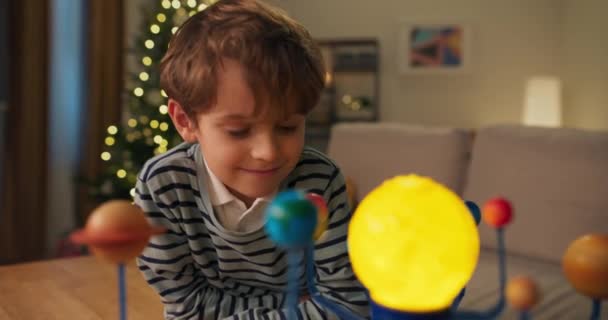 太陽系の子供モデルを見て笑顔ハンサムな男の子 彼はシャツとストライプのセーターを着ている 少年は惑星を研究し クリスマスツリーが後ろにちらつき — ストック動画