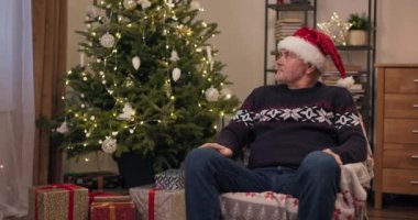 Odada Noel ağacı var, çelenkler titreşiyor ve hediyeler var. Yalnız, orta yaşlı bir adam sandalyeye oturur, kafasını çevirir ve kameraya bakar. Sıcak bir tatil süveteri ve Noel Baba şapkası giyiyor..