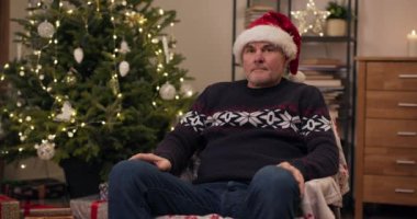 Emekli bir adam sandalyede oturur ve başka tarafa bakar. O, sıcak tatil kazağı ve kafasına Noel Baba şapkası giyiyor. Arkasında titreyen çelenklerle Noel ağacı var. Adam üzgün ve ciddi bir şekilde kameraya bakıyor..