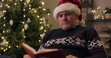 Emekli bir adam Noel ağacının yanında oturmuş elinde çelenklerle titreyerek kitap tutuyor. O, dikkatle roman okuyor ve sayfayı çeviriyor. Adam kalın bir kazak giymiş ve Noel Baba şapkası takıyor.