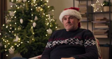 Noel ağacının yanındaki sandalyede oturan gri saçlı emekli adam. O, sıcak tatil kazağı ve Noel Baba şapkası giyiyor. Adam gergin bir şekilde şapkasını çıkardı, çünkü kutlama havasında değildi..