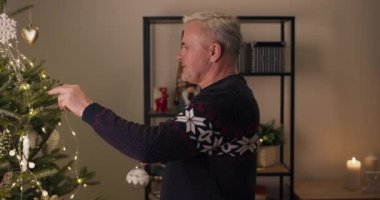 Mutlu büyükbaba Noel ağacını süslüyor. Dekorasyonu tutuyor, asıyor ve çelenkleri tamir ediyor. Adam süveter giymiş ve Noel 'i kutlamak için çocukları ve torunlarını bekliyor..