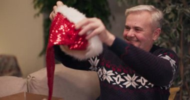 Gri saçlı neşeli orta yaşlı adam Noel Baba şapkasını kutusundan çıkarıp kafasına geçirir ve güler. O, sıcak tatil kazağı giyiyor. Şimdi yeni yılı kutlamaya hazır..