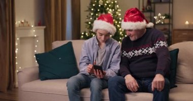 Mutlu baba ve oğlu Noel Baba şapkası takmış oturma odasında kanepede oturuyor. Çocuk telefonu elinde tutuyor ve babasına çektiği komik videoyu gösteriyor. Babam telefona bakar ve yüksek sesle güler..